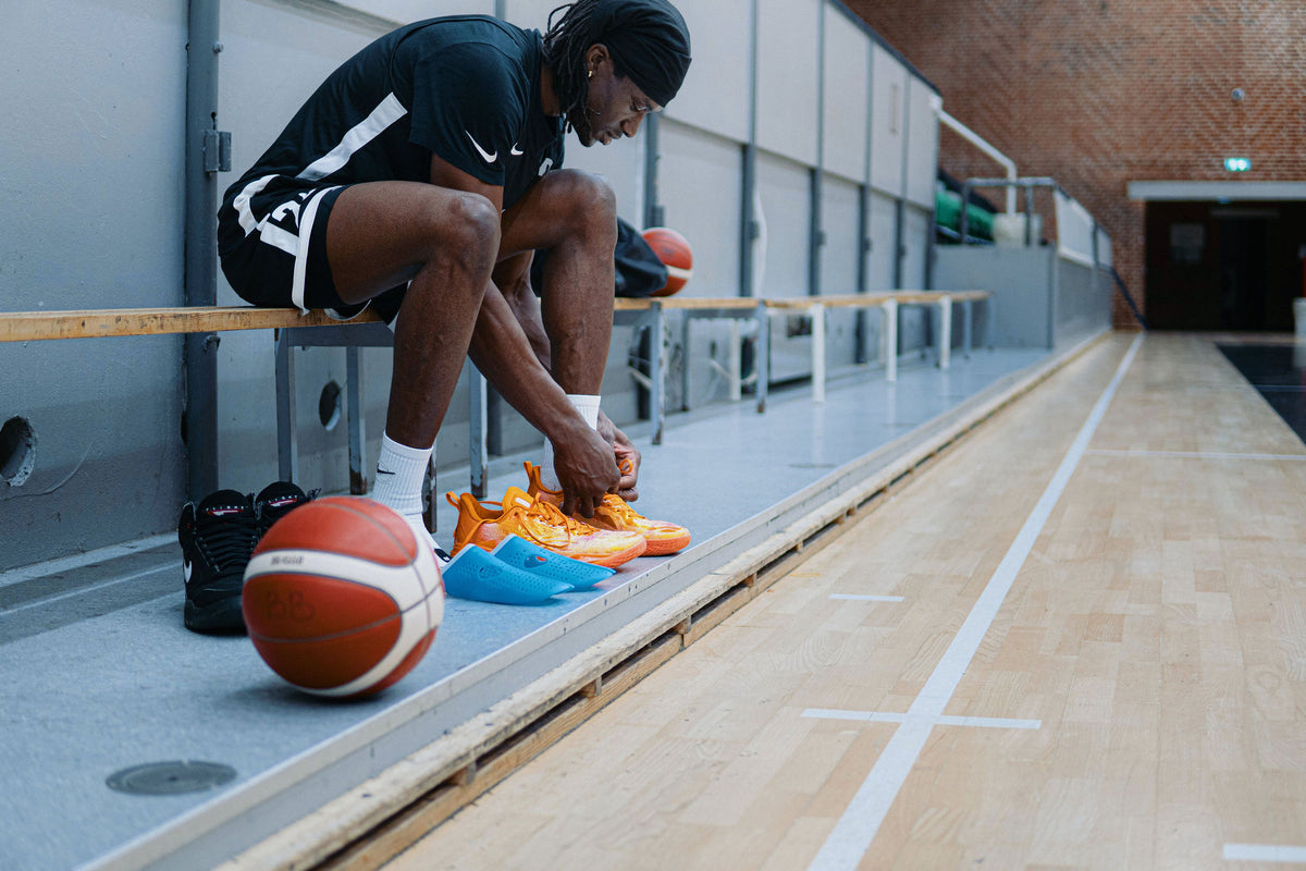 An athlete on a courtside bench unlacing orange basketball shoes, with two blue SHAPES ready beside them
